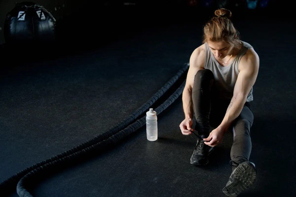 Deportista sentado en el suelo de un gimnasio atándose las zapatillas junto a una botella y cuerda de batalla