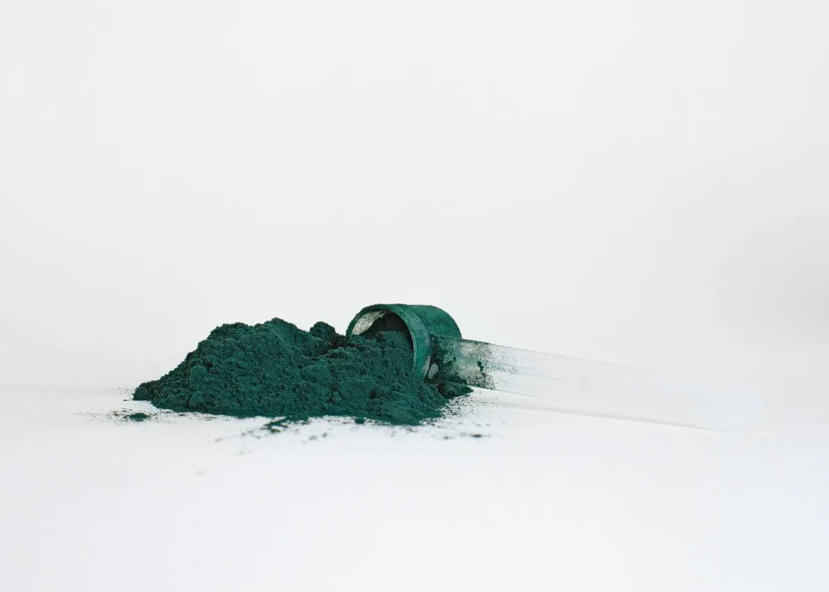 Montículo de polvo verde de proteína vegetal con cuchara medidora sobre fondo blanco.png